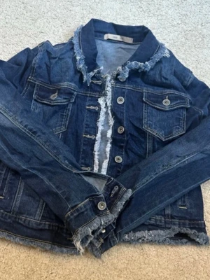 Mörkblå jeansjacka med slitna detaljer - Snygg mörkblå jeansjacka med fransade och slitna kanter vid krage, knappslå och nederkant. Klassisk modell med bröstfickor och silverfärgade knappar. Perfekt för dig som gillar en lite mer edgy och avslappnad stil. Från Costa Mani