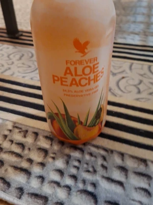Forever Aloe Peaches dryck - En flaska Forever Aloe Peaches dryck med 84,5% aloe vera gel och persikosmak. Flaskan är vit och orange med tryck i orange och grönt, och har en bild på aloe vera-blad och persikor på etiketten. Perfekt för dig som gillar fräscha smaker.