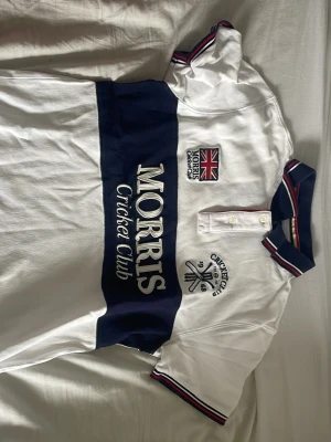 Vit och marinblå cricketpolo Morris - Snygg vit pikétröja från Morris Cricket Club med bred marinblå rand över bröstet, broderade detaljer och brittisk flagga. Kragen har röd, vit och marinblå rand. Klassisk knappslå framtill och korta ärmar med randiga muddar.