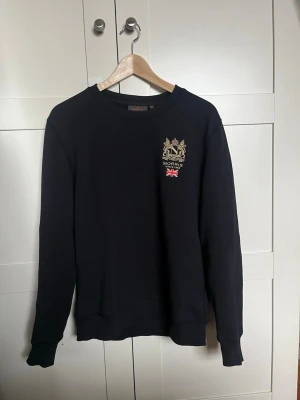 Svart sweatshirt från Morris - Svart sweatshirt från Morris med rund halsringning och broderad logga samt brittisk flagga på bröstet. Tröjan har långa ärmar och ribbade muddar vid ärmslut och nederkant. Perfekt för en avslappnad och stilren look.