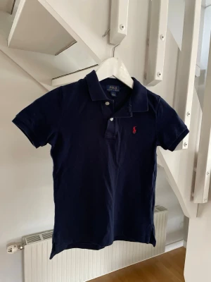 Marinblå pikétröja från Polo Ralph Lauren - Klassisk marinblå pikétröja från Polo Ralph Lauren med korta ärmar och krage. Tröjan har två knappar framtill och den ikoniska röda logotypen broderad på bröstet. Tillverkad i mjuk bomull som känns skön mot huden.