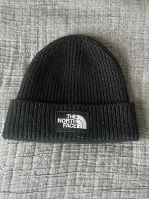 Svart ribbstickad mössa The North Face - Säljer en svart ribbstickad mössa från The North Face med uppvikt kant och tydlig logotyp framtill. Klassisk modell i mjukt material som passar perfekt till kyliga dagar. Enkel och stilren design som funkar till det mesta.
