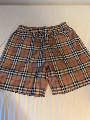 Rutig Burberry shorts med resårmidja - Säljer ett par klassiska Burberry shorts med det ikoniska rutiga mönstret i beige, svart, vitt och rött. Shortsen har resår i midjan för skön passform och är perfekta för sommaren. Styla med en enkel t-shirt för en clean look. Passar M