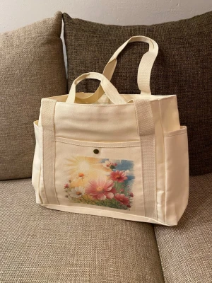 Cremefärgad väska/ shoppingväska/strandväska med blommotiv - Snygg beige shoppingväska i canvas med tryckt blommotiv i rosa, vitt och grönt på framsidan. Väskan har två långa handtag, en ytterficka med knapp och sidofickor för extra förvaring. Perfekt för dig som vill ha en unik och praktisk väska.