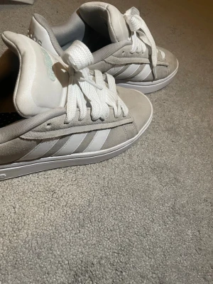 Gråa Adidas sneakers med vita ränder - Snygga gråa Adidas sneakers med klassiska vita ränder på sidorna och vita skosnören. Skorna har en låg profil och är tillverkade i mockaliknande material med textilfoder för extra komfort. Perfekta för dig som gillar stilrena och tidlösa sneakers. Lite smutsig lätt ta bort av tvätt och köpte för 1000kr