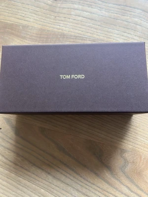 Tom Ford solglasögon med brun ask - Säljer ett par Tom Ford solglasögon i originalförpackning.  Medföljer en lyxig brun sammetsfodral med guldfärgad logga. Perfekt för dig som vill ha en exklusiv accessoar från ett av världens mest ikoniska modemärken. Orginal pris 2600 om du är osäker så kan du åka till synsam för att testa  dem.