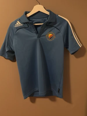 Blå DIF piké från Adidas - Snygg ljusblå piké från Adidas med Djurgårdens IF-logga på bröstet. Tröjan har klassiska vita Adidas-ränder på axlarna, korta ärmar och krage med knappar. Tillverkad i funktionsmaterial.