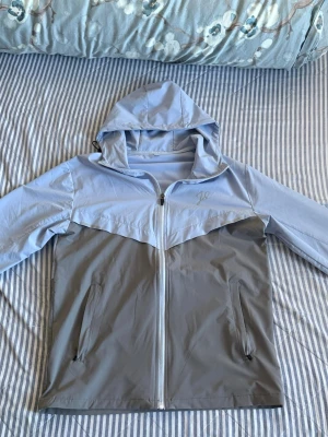 Tvåfärgad vindjacka med huva - Snygg vindjacka i ljusblått och grått med dragkedja framtill och två fickor med dragkedja. Jackan har huva och elastiska muddar vid ärmsluten. Perfekt för blåsiga dagar och enkel att matcha till olika outfits.