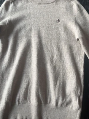 Beige stickad tröja Morris - Snygg beige stickad tröja från Morris med rund halsringning och diskret broderad logga på bröstet. Tröjan har ribbade muddar vid ärmslut och nederkant. Perfekt för lager på lager och enkel att matcha.