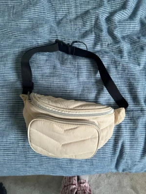 Beige quiltad magväska - Snygg beige magväska med quiltad design och två dragkedjefack. Väskan har justerbart svart band och silvriga dragkedjor. Perfekt för att bära det viktigaste och ger en trendig touch till din outfit.