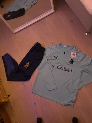 Olympique Marseille träningsset Puma - Snyggt träningsset från Olympique Marseille och Puma. Tröjan är ljusblå med halv dragkedja, klubbmärke och sponsortryck. Byxorna är mörkblå med klubbmärke och har smal passform. Perfekt för fotbollsträning eller chill. Materialet är funktionspolyester.