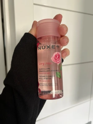 Nuxe Very Rose micellärvatten - Nuxe Very Rose Soothing Cleansing Micellar Water med rosextrakt. Kommer i en genomskinlig plastflaska med rosa lock och etikett med rosdesign. Innehållet är ljusrosa och flaskan rymmer 100 ml. Perfekt för att ta bort smink och rengöra huden.