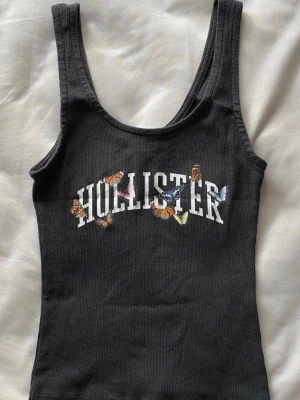 Vintage hollister topp - Snygg svart ribbad linnetopp från Hollister med broderade fjärilar och stor logga framtill. Toppen har breda axelband och är figurnära, perfekt för en trendig och avslappnad look.