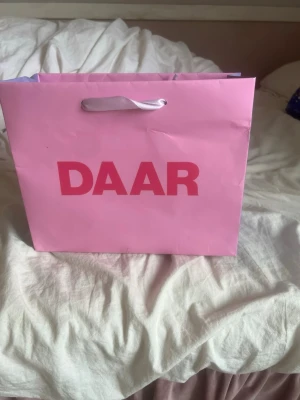 Rosa shoppingväska från DAAR - Snygg shoppingväska i rosa papper från DAAR med breda handtag i vitt band. Väskan har en rektangulär form och tryckt logga framtill. Perfekt för att bära med sig grejer eller använda som presentpåse.