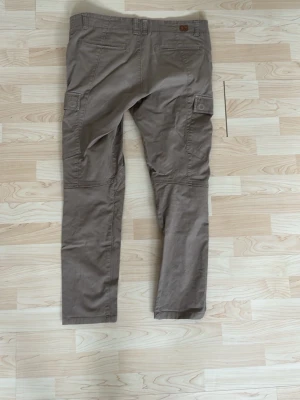 Beiga boomerang cargobyxor med fickor - Säljer ett par beiga cargobyxor med raka ben och stora sidofickor med lock. Byxorna har bälteshällor och detaljerade sömmar vid knäna för extra stil. Perfekta för dig som gillar en avslappnad och funktionell look.