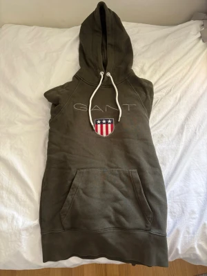 Olivgrön hoodieklänning från GANT - Snygg olivgrön hoodieklänning från GANT med stor huva och vita dragsnören. Framtill finns en känguruficka och ett broderat GANT-emblem med amerikansk sköld. Tillverkad i mjuk bomullsblandning, perfekt för en avslappnad look.