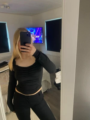 Svart långärmad topp med rynk - Trendig svart långärmad topp med rynkade detaljer framtill och lätt croppad passform. Toppen har en fyrkantig ringning och är tillverkad i ett glansigt, stretchigt material som ger en snygg siluett. 