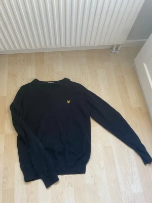 Svart tröja från Lyle & Scott - Svart stickad tröja från Lyle & Scott med klassisk rund halsringning och gul logotyp på bröstet. Tröjan har långa ärmar och ribbade muddar vid ärmslut och nederkant. Perfekt för dig som gillar stilrena plagg med diskret branding.