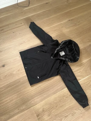 Svart vindjacka från Moncler - Snygg svart vindjacka från Moncler med huva och dragkedja. Jackan har logotyp på bröstet och Moncler-tryck på huvkanten. Två sidofickor med dragkedja och elastiska muddar vid ärmsluten. Perfekt för blåsiga dagar.