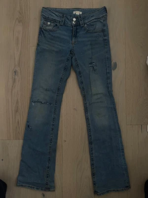 Blå slitna bootcut jeans - Ett par blå bootcut jeans med slitningar och råa detaljer på benen. Jeansen har klassisk femficksdesign, knappgylf och bälteshällor. Materialet är denim i bomull med en tvättad look som ger en avslappnad vibe.