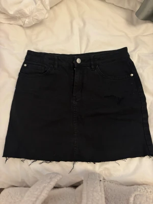 Svart jeanskjol med rå kant - Säljer en svart jeanskjol med fem fickor och rå, fransig kant nedtill. Kjolen har knapp och dragkedja framtill samt bälteshällor. Perfekt basplagg till garderoben! Jag har klippt kjolen kortare själv, så den är lite kortare där bak än fram. Har även klippt ett litet hål där fram som mode. 