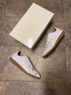 Maison Margiela Paint Splatter Gats - Unika vita sneakers från Maison Margiela med färgstänk i rött, blått, gult och svart. Skorna har beige sula och vita snören. Tillverkade i läder med klassisk låg siluett och diskreta sömmar. Perfekta för dig som vill sticka ut med din stil. True to size