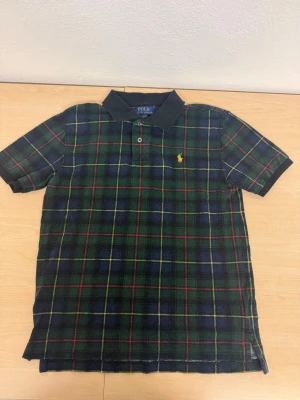 Rutig pikétröja från Polo Ralph Lauren - Klassisk pikétröja från Polo Ralph Lauren med rutigt mönster i mörkblått, grönt, rött och gult. Tröjan har korta ärmar, krage och knappslå framtill samt den ikoniska broderade loggan på bröstet. Perfekt för en avslappnad men stilsäker look.