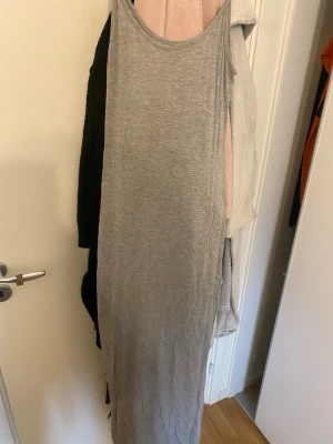 Grå långklänning i bomull - En enkel och stilren grå långklänning i mjukt bomullsmaterial. Klänningen är ärmlös med smala axelband och har en rak, kroppsnära passform som ger en clean look. Perfekt att styla med en kofta eller jacka. Använd några gånger men inga slitningar osv fortfarande fint skick. 