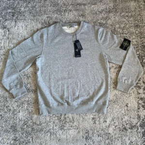 Grå stickad tröja från Stone Island - Snygg grå stickad tröja från Stone Island med rund halsringning och ribbade muddar. Tröjan har den klassiska Stone Island-loggan på vänster ärm och är tillverkad i mjukt material för en avslappnad look.