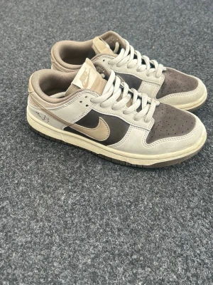 Nike Dunk Low i beige och brunt - Säljer ett par Nike Dunk Low sneakers i beige och brunt med klassisk swoosh på sidan. Skorna har en låg siluett, perforerad tåbox och kontrasterande paneler i mocka och läder. Perfekta för dig som gillar streetwear och vill ha en stilren look.