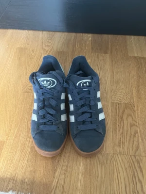 Mörkblå Adidas sneakers med gummisula - Snygga mörkblå sneakers från Adidas med klassiska vita ränder på sidorna och brun gummisula. Skorna har låga skaft och är tillverkade i mockaliknande material. Perfekta för dig som gillar en sportig och stilren look.