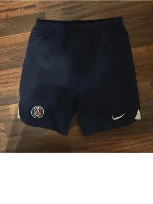PSG Nike Dri-Fit shorts marinblå - Marinblå träningsshorts från Nike med PSG-logga på ena benet och vit Nike Swoosh på det andra. Tillverkade i Dri-Fit-material som andas och transporterar bort fukt. Perfekta för fotboll eller annan träning.