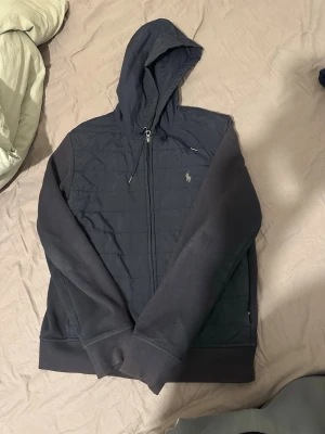  Polo Ralph Lauren Jacka - Snygg mörkblå hoodie från Polo Ralph Lauren med dragkedja framtill och broderad logga på bröstet. Huva med dragsko och quiltad front i glansigt material, medan ärmar och rygg är i mjuk bomull. Perfekt för en chill och stilren look.