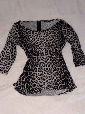 Leopardmönstrad topp/blus med trekvartsärm - Snygg topp med leopardmönster i svart och beige. Modellen har trekvartsärm, rundad halsringning och dragkedja i ryggen. Materialet är stretchigt och följsamt, perfekt för dig som vill sticka ut lite extra.