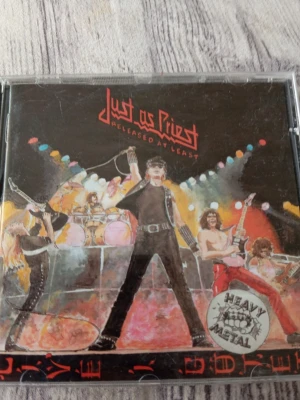 Judas  Priest CD - Released at Least - CD-skiva med albumet 'Released at Least' från Judas  Priest. Omslaget har en färgstark illustration av ett heavy metal-band på scen med starka ljus och instrument. Skivan är svart med röda cirklar och text. Perfekt för dig som gillar hårdrock och samlar på musik.