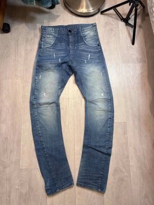 Blå slitna jeans med smal passform - Säljer ett par blå jeans med smal passform och slitna detaljer på både fram- och baksida. Jeansen har klassisk femficksdesign, ljusa tvättade partier och subtila slitningar för en trendig look. Perfekta för dig som gillar en avslappnad men stilsäker stil.