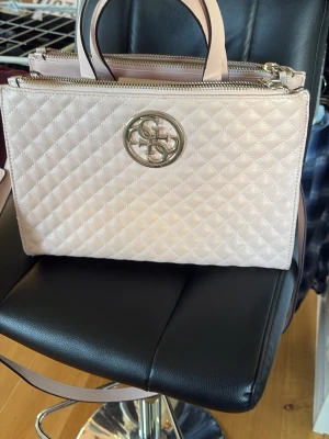 Ljusrosa quiltad handväska Guess - Snygg ljusrosa handväska från Guess med quiltat mönster och guldfärgad logga framtill. Väskan har både handtag och axelrem samt dragkedja upptill. Tillverkad i konstläder och levereras med dustbag från Guess.
