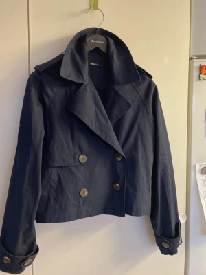 Mörkblå kort trenchcoat - Kort trenchcoat i mörkblått med dubbelknäppning och stora knappar. Jackan har bred krage, axelklaffar och dekorativa remmar vid ärmsluten. Tillverkad i ett slätt tyg som ger en stilren look. Perfekt för dig som vill ha en klassisk men modern jacka.