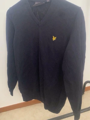 Svart tröja med V-ringning Lyle & Scott - Klassisk svart tröja från Lyle & Scott med V-ringning och den ikoniska gula örnloggan broderad på bröstet. Tröjan har långa ärmar och ribbade muddar vid ärmslut och nederkant. Perfekt för en stilren och enkel look.