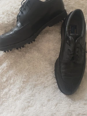 Svarta golfskor från FootJoy - Säljer ett par svarta golfskor från FootJoy med klassisk snörning och grov sula för bra grepp på banan. Skorna är tillverkade i läder med en stilren design och diskreta detaljer. Perfekta för dig som vill ha både komfort och stil på golfbanan.