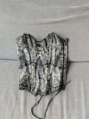 Svart spetskorsett Victorias secret - Säljer en snygg korsett i svart spets med transparent mönster och dekorativ rosett framtill. Korsetten har snörning bak och markerade sömmar för en klassisk siluett. Materialet är en kombination av mesh och spets som ger en elegant look.