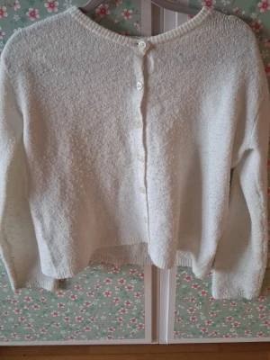 Vit stickad kofta med knappar - Mysig vit stickad kofta med rund halsringning och knäppning framtill. Koftan har långa ärmar och ribbstickade muddar vid ärmslut och nederkant. Perfekt att slänga på sig när det är kyligt ute.