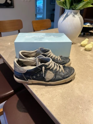 Philippe Model blå låga sneakers - Snygga blå sneakers från Philippe Model med vita detaljer och gråa skosnören. Skorna har en låg siluett och är tillverkade i läder med gummisula. Klassisk design med logotyp på sidan och kontrastfärgade partier vid häl och tå. Skorna har defekter därav priset men några utav defekterna är design som ska vara där ( de som är vid sulan för det mesta)