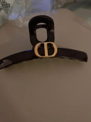 Svart hårklämma med dior detalj  - Snygg svart hårklämma från Dior med en elegant guldfärgad logga framtill. Klämman är tillverkad i glansig plast och har en klassisk, böjd form som håller håret på plats. Perfekt accessoar för att lyfta din look.