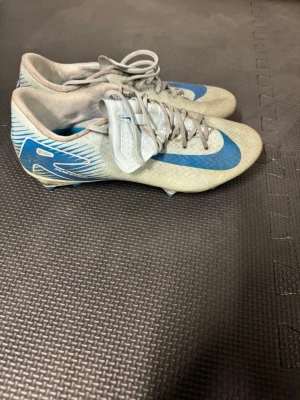 Nike Mercurial blå/vita fotbollsskor - Säljer ett par Nike Mercurial fotbollsskor i vitt och blått med klassisk Nike-swoosh på sidan. Skorna har snörning och låg profil, tillverkade i syntetmaterial för bra grepp och känsla på planen. Perfekta för dig som vill ha lätta och snabba skor till match eller träning.