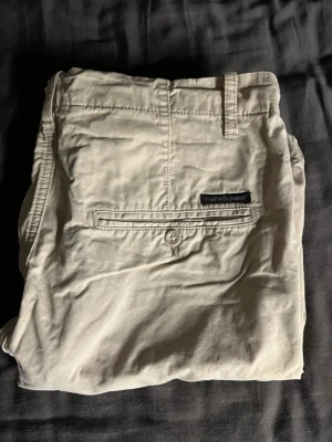 Beige shorts från Peak Performance - Snygga beige shorts från Peak Performance med klassisk passform. Shortsen har bälteshällor, två bakfickor med knapp och diskret logga ovanför ena fickan. Perfekta för sommardagar och enkla att matcha med olika stilar.