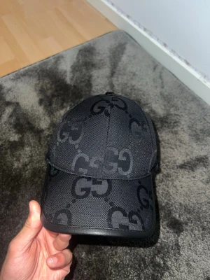 Svart keps med GG-mönster - Snygg svart keps med det klassiska GG-mönstret över hela kepsen. Kepsen har böjd skärm och är tillverkad i ett tåligt tygmaterial. Perfekt accessoar för dig som vill ha en stilren och exklusiv look.