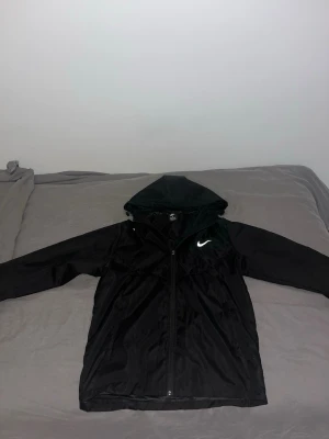 Svart Nike vindjacka  - Snygg svart vindjacka från Nike med reflektiv logga på bröstet. Jackan har dragkedja framtill, huva och långa ärmar. Tillverkad i lätt och tåligt material som skyddar mot vind. Perfekt för dig som vill ha en sportig och stilren look. Det står storlek M men den passar storlek S.