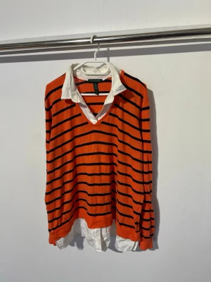 Randig orange långärmad topp - Säljer en orange långärmad topp från Ralph Lauren med svarta horisontella ränder och vit skjortkrage. Toppen har v-ringning och en lös passform, samt en vit skjortdetalj som sticker ut nedtill och vid kragen. Perfekt för en trendig lager-på-lager-look.
