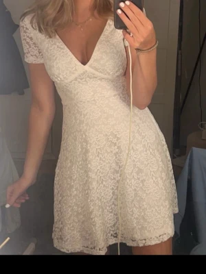 Vit studentklänning/spetsklänning - Supersöt vit kortklänning i spets med v-ringad hals och korta ärmar. Klänningen har en omlott-look upptill och ett fint blommigt spetsmönster över hela tyget. Perfekt för sommarens alla tillfällen!